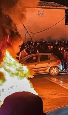 Fogo na rua. Ao redor há várias pessoas e um carro. 