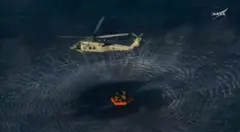 Um helicóptero da Marinha sobrevoa uma balsa que flutua no mar e transporta a tripulação da Artemis II. A força de suas hélices cria ondas concêntricas na água