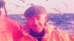 Old photo of fisherman on a fishing boat, wearing waterproof oils and a cap, the man - Jim Runcie - also has a cigarette in his mouth, sea and lots of seagulls in the background.