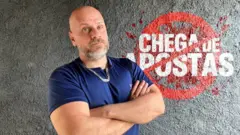 Rafael Santos, um homem corpulento, careca, de barba grisalha e olhos azuis, com os braços cruzados e uma expressão séria no rosto, a frente de um logotipo onde se lê: "Chega de apostas"