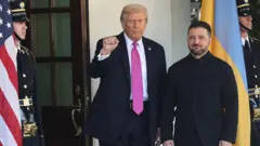 ډونلډ ټرمپ له زيلېنسکي سره ولاړ دی او ښی لاس یې موټی او اوچت کړی دی.