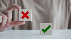 Mão de mulher segurando um bloco com um X vermelho, enquanto outro bloco com um tique verde repousa sobre a mesa