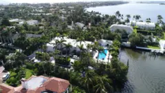 Vista aérea de la casa de Jeffrey Epstein en Palm Beach, que muestra una gran casa blanca en un barrio costero, con una piscina visible entre numerosas palmeras.