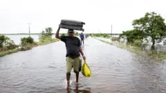 Um homem caminha por uma estrada inundada com uma mala na cabeça.