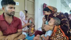 অক্সিজেন মাস্ক লাগানো দুটি শিশু কোলে নিয়ে একটি বিছানার ওপর বসে আছেন দুই জন নারী। পাশে একজন পুরুষ