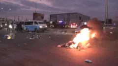 A pile of rubbish burns on the highway at dusk as traffic tries to get past.