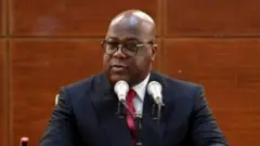 Perezida Félix Tshisekedi wa RDC arimo kuvuga ijambo yicaye imbere y'indangururamajwi ebyiri, inyuma ye hari urukuta rw'ibara ry'ikigina. Yambaye ikoti ry'umukara n'ishati y'umweru na karuvati ('cravate') y'umutuku ujya kuba iroza, n'indorerwamo. Ifoto yo ku wa gatandatu, i Kinshasa, ku itariki ya 18 Mutarama (1) mu mwaka wa 2025. 