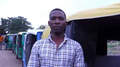 Un homme portant une chemise à carreaux se tient devant son taxi à trois roues.
