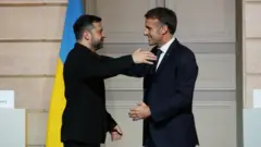 クリーム色の石の壁や扉、ウクライナの旗を背に、黒い上着のゼレンスキー氏とダークスーツのマクロン氏が共に笑顔を浮かべ、両手を広げて近づいている