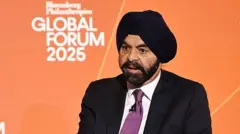 Ajay Banga discursando em frente a um fundo laranja, vestindo um turbante preto, terno preto e gravata roxa.