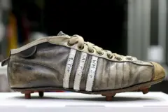 Botín de fútbol con tacos, de color negro desteñido, las tres franjas de Adidas en blanco con algo escrito en ellas.