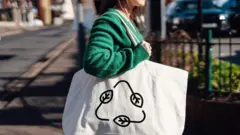 Mulher anda na rua com ecobag com estampa que ilustra símbolo da reciclagem