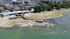 Imagem aérea do sítio arqueológico de Iznik, com alvenaria parcialmente submersa, delineando as antigas paredes de uma igreja. Um mirante se estende até as águas a partir de um edifício próximo.
