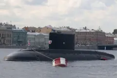 Черная подводная лодка частично погружена в воду, с рубкой и флагом на корме. Она стоит у красно-белого буя на реке. На заднем плане — ряд исторических зданий в светлых и пастельных тонах вдоль набережной под пасмурным небом.