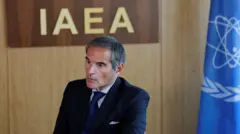 Рафаэль Гросси сидит в костюме за столом на фоне букв IAEA, сбоку от него виден флаг ООН.