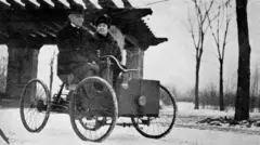 A imagem em preto e branco mostra duas pessoas sentadas em um veículo antigo de quatro rodas finas e estrutura simples, semelhante aos primeiros automóveis. O homem conduz o veículo enquanto a mulher está sentada ao lado dele, ambos usando roupas pesadas e chapéus típicos do início do século 20. Ao fundo, aparecem árvores sem folhas e uma construção com telhado inclinado em uma paisagem de inverno.