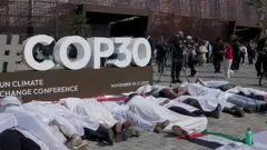 Manifestantes acostados frente a un aviso de la COP30, cubiertos en sábanas blancas.