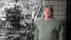 Nikolai Solovyov, engenheiro-chefe da sala de turbinas no momento do acidente, aparece na foto em frente a uma imagem do reator destruído, no museu Slavutych. Ele tem cabelos grisalhos, veste um moletom verde-acinzentado e olha para a câmera com uma expressão séria.