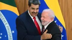 A imagem mostra dois homens se abraçando e sorrindo durante um encontro oficial, tendo ao fundo bandeiras do Brasil e da Venezuela. Um deles segura uma caneta na mão enquanto ambos posam em um gesto amistoso diante das câmeras.