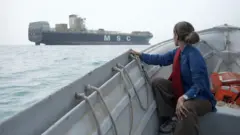 一名女子坐在一艘灰色小船上,望著海面上的一艘大型貨輪,船身側面印有白色的「MSC」字樣。她留著深色長髮,將其束在腦後,身穿一件藍色上衣。她身後放著幾件橘色的救生衣。