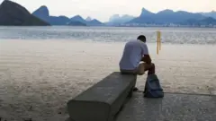 Homem olhando para baixo e sentado em banco na beira de praia em Niterói, com vista do Rio de Janeiro ao fundo