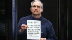 Paul Whelan holds up a sign at the end of his trial - "Sham trial! Meatball surgery! No human rights! Paul's life matters! Decisive action from potus and PMs needed! Happy Birthday Flora!"