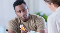 Un hombre sostiene un frasco de pastillas mientras mira con preocupación a una doctora