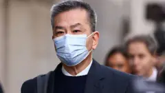 袁松彪抵达伦敦中央刑事法院。他留有短灰发，佩戴蓝色外科口罩遮住脸部下半部，身穿深色外套，内搭黑色针织上衣与白色有领衬衫