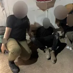 Foto de um homem e um jovem em um quarto, um deles com roupas militares. Os rostos estão borrados.