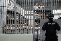 Presos com uniformes brancos estão na cela de uma prisão enquanto são observados por um policial 