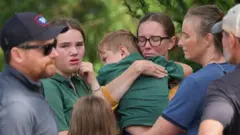 চার-পাঁচ বছরের একটি শিশু কোলে চোখ বন্ধ করে আছেন এক নারী, তার সামনে একটি ছোটো মেয়ে। পাশে আরো চার জন মানুষ 