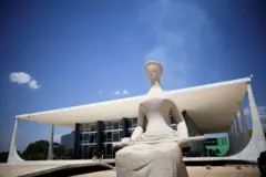 Estátua de pedra da Justiça em primeiro plano, sentada e segurando uma espada, em frente ao prédio do Supremo Tribunal Federal, em Brasília, sob céu azul intenso.