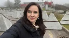 Премьер-министр Литвы Инга Ругинине в черной куртке делает селфи на фоне пограничных ограждений, за которыми начинается Беларусь