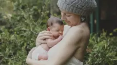 一位戴帽子的女子（考特尼·路易絲）在戶外，抱著她剛出生的孩子，孩子貼著她因懷孕而隆起的腹部。 