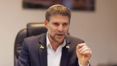 بتسلئيل سموتريتش، وزير المالية الإسرائيلي، يرتدي بدلة زرقاء داكنة، وقميصًا أبيض، وكيباه أسود. يرفع يده اليسرى مع توجيه إصبعه السبابة إلى الأعلى.