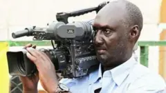 Valence Rwamukwaya arimo gufata amashusho na 'camera' nini iri ku rutugu. 