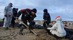 Люди сгребают лопатой в черные мешки загрязненный песок. Двое из них в защитных костюмах.