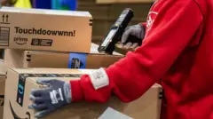 赤いプルオーバーを着たアマゾンの従業員が、配送用の段ボール箱を手に持った端末でスキャンしている。従業員は上半身の肩から下だけが写っている