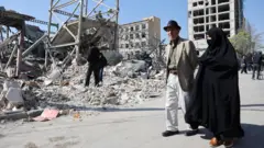 Tahran'da yıkılan binalar arasında yürüyen şapkalı bir erkek ve yanında siyah çarşaflı bir kadın