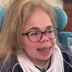 Sophia Scott sonríe ampliamente, mirando fuera de cámara. Tiene el cabello largo y rubio, y lleva gafas de montura marrón. Viste una blusa azul con ribetes rosas y una bufanda de algodón de color rosa pálido alrededor del cuello.