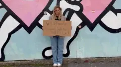 Barbora is standing in front of a wall mural on Tyneside and holding a sign that reads "selfcare tip: regularly check your boobs." She has long blonde hair and is wearing jeans and trainers. 