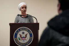  Ilhan Omar, kürsüde konuşma yapıyor, sırtı dönük bir erkek ekran önünde görünüyor
