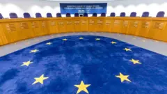 Yerde üzerinde yıldızlar olan bir halı. Arkada kürsü ve Avrupa İnsan Hakları Mahkemesi yazısı