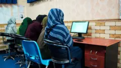 تصویر چهار دختر در یک صنف آموزش کمپیوتر. دختران در عقب میز و پشت شان به سمت دوربین نشسته اند. در مقابل شان یک کمپوتر دیده می‌شود. یک معلم زن هم در تصویر دیده می شود که به دختران رهنمایی می‌کند. 