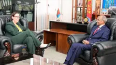 ማሪያ ማልመር ስተነርጋረድ ምስ ኤርትራዊ መዘናኣ ዑስማን ሳልሕ ኣብ ቤት ጽሕፈቱ ኮፊ ኢሎም እናተዘራረቡ፡ ባንዴራ ኤርትራን ፋይላትን ኣብ ድሕረባይታኦም ይርአ።