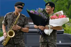 O integrante do grupo de K-pop BTS, RM (à esquerda), toca saxofone enquanto V observa segurando buquês de flores, em um espaço esportivo ao ar livre em Chuncheon, em 10 de junho de 2025