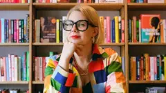 A escritora Emma Gannon, de óculos e roupa colorida, sentada em frente a uma estante de livros