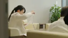 La alcaldesa Shoko Kawata, una mujer sentada en un sillón beige, revisa varias páginas impresas en una sala de reuniones.