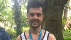 Will veste uma camiseta esportiva e usa uma faixa azul ao redor do pescoço, que parece ser uma medalha. Ele sorri para a câmera e está em um ambiente cheio de árvores.