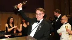 Улыбающийся Павел Таланкин в смокинге держит статуэтку «Оскара» на фоне нарядно одетых людей 